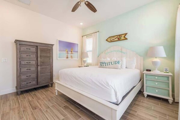A bedroom with a bed, a dresser, a nightstand, a ceiling fan, a window with curtains, and a wall-mounted sign.