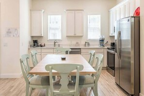 A kitchen with a dining table, chairs, cabinets, a refrigerator, and a coffee maker.