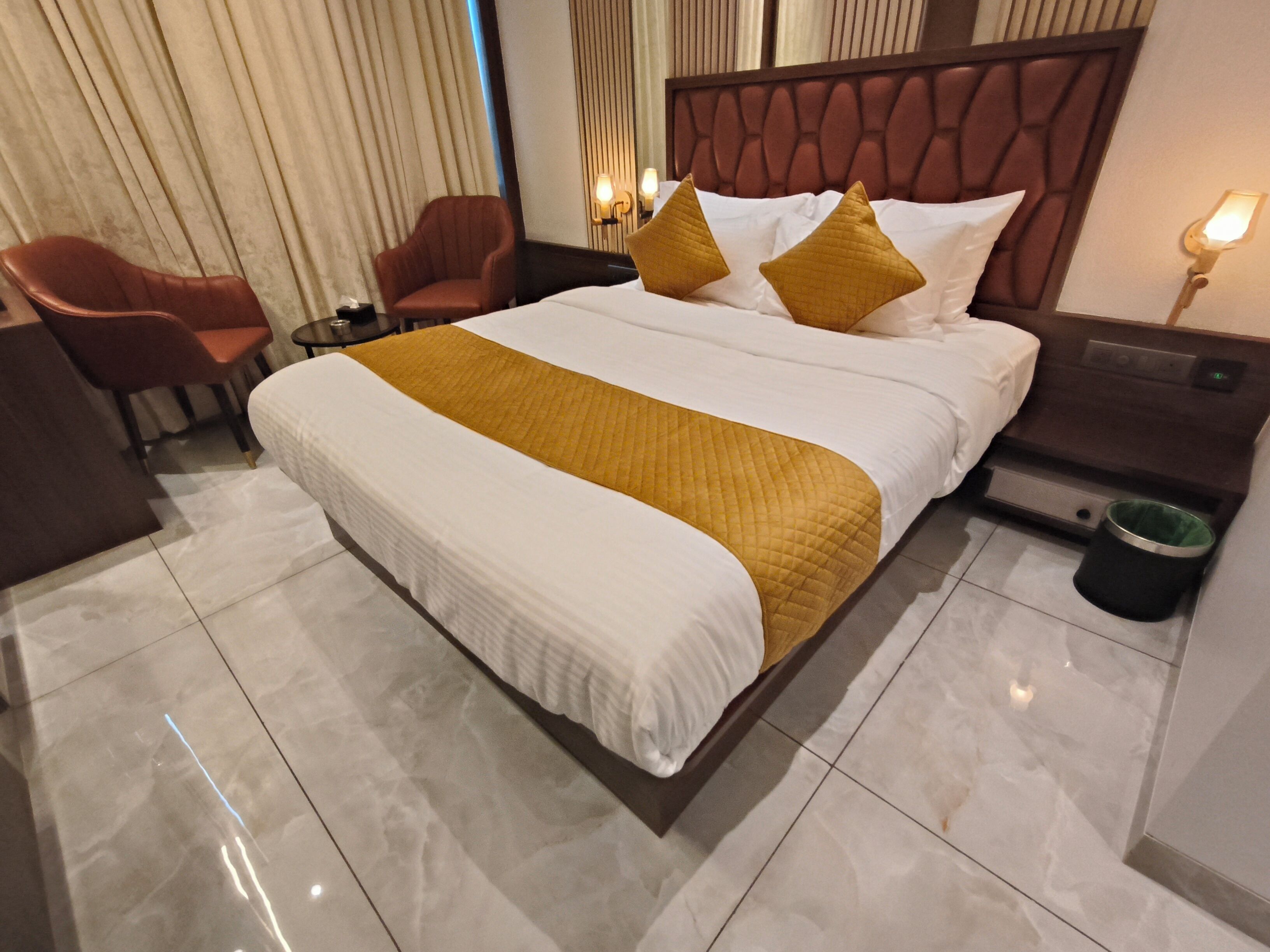 A hotel room with a large bed, a wooden headboard, a nightstand with a lamp, a chair, and a trash bin.