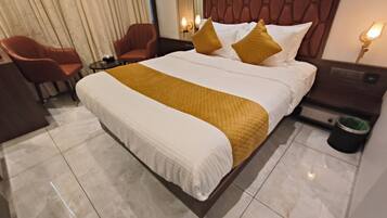 A hotel room with a large bed, a wooden headboard, a nightstand with a lamp, a chair, and a trash bin.