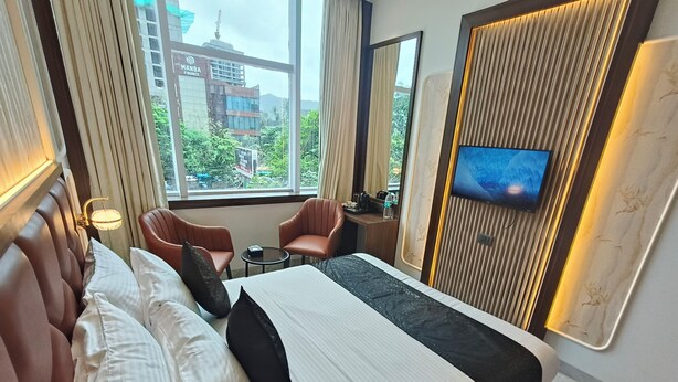 A hotel room with a large bed, two armchairs, a small table, a TV mounted on the wall, and a view of buildings outside the window.