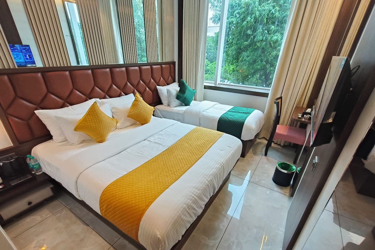 Ein Hotelzimmer mit einem großen Bett, zwei kleineren Betten, einem Fernseher, einer Kaffeemaschine und Blick auf Bäume durch das Fenster.