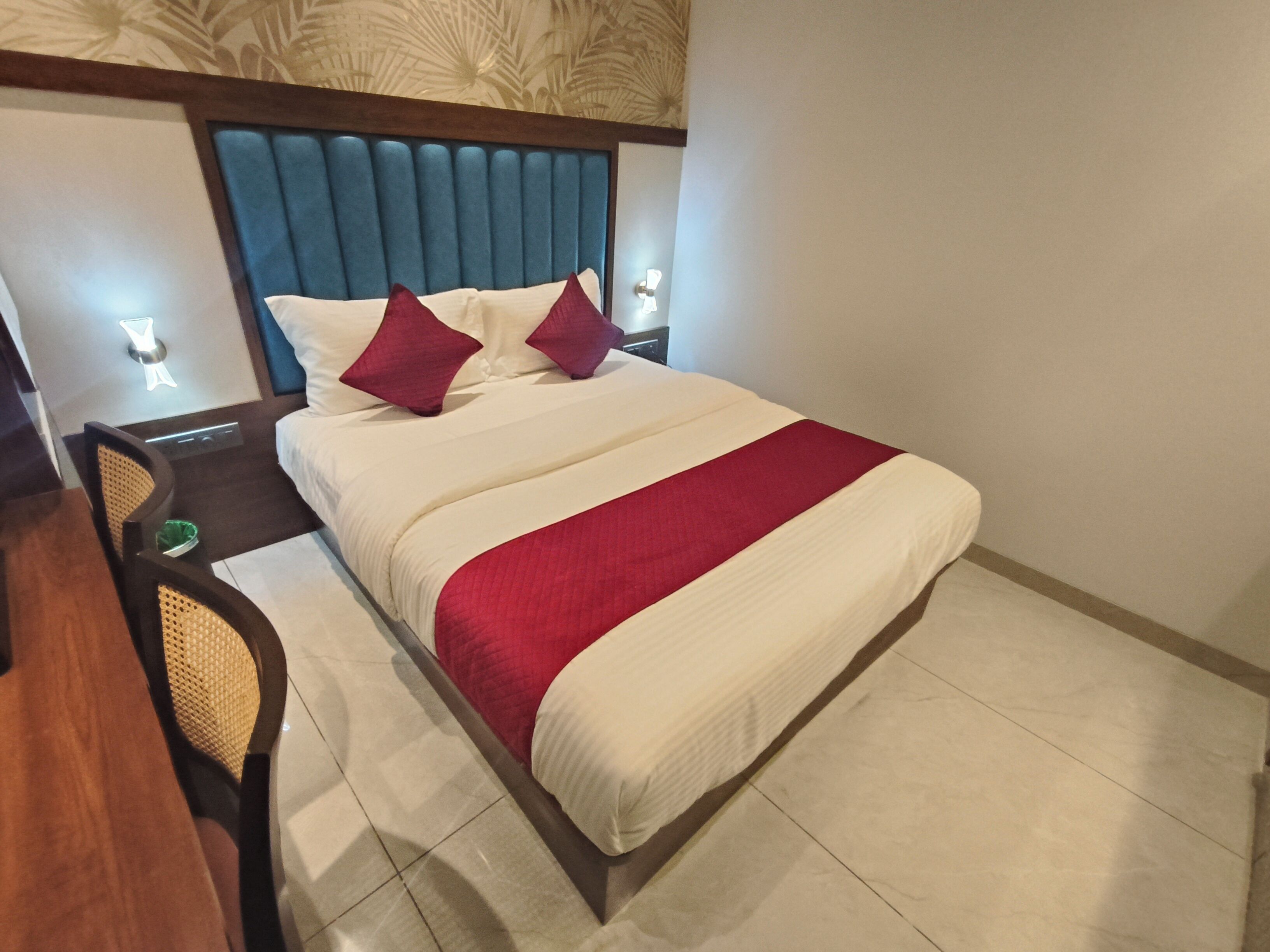 Ein Hotelzimmer mit einem großen Bett, zwei Kissen, einer roten und weißen Tagesdecke, einem Holzkopfteil, einem Nachttisch mit Lampe und einem Stuhl mit gewebter Sitzfläche.