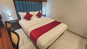 Habitación de hotel con una cama grande, dos almohadas, una colcha roja y blanca, cabecera de madera, mesita de noche con lámpara y una silla con asiento de mimbre.