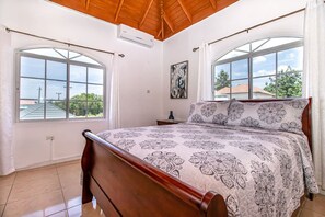 A bedroom with a wooden bed, two windows with curtains, a painting on the wall, and a tiled floor.