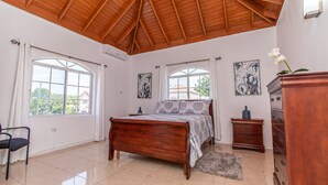 A bedroom with a wooden ceiling, a bed with floral bedding, two windows, a dresser, and a chair.