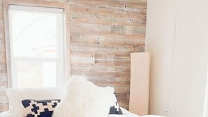 Un dormitorio con pared de madera, cama con ropa de cama blanca, banqueta con cojines estampados y una lámpara colgante.