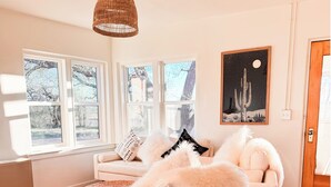 Una sala de estar con un sofá blanco, una lámpara colgante tejida y una obra de arte con temática de cactus en la pared.