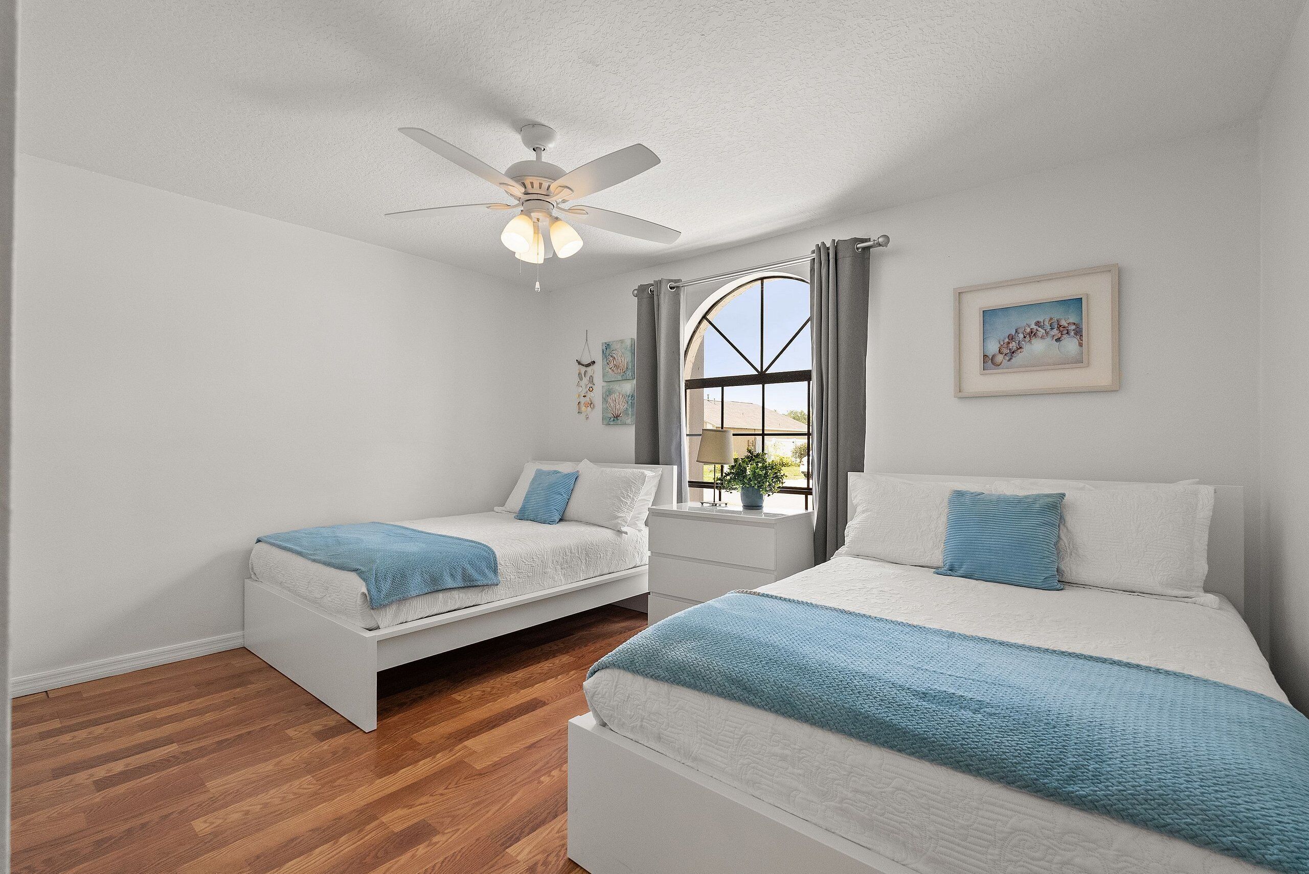 A bedroom with two beds, a ceiling fan, a window with curtains, and wall art.