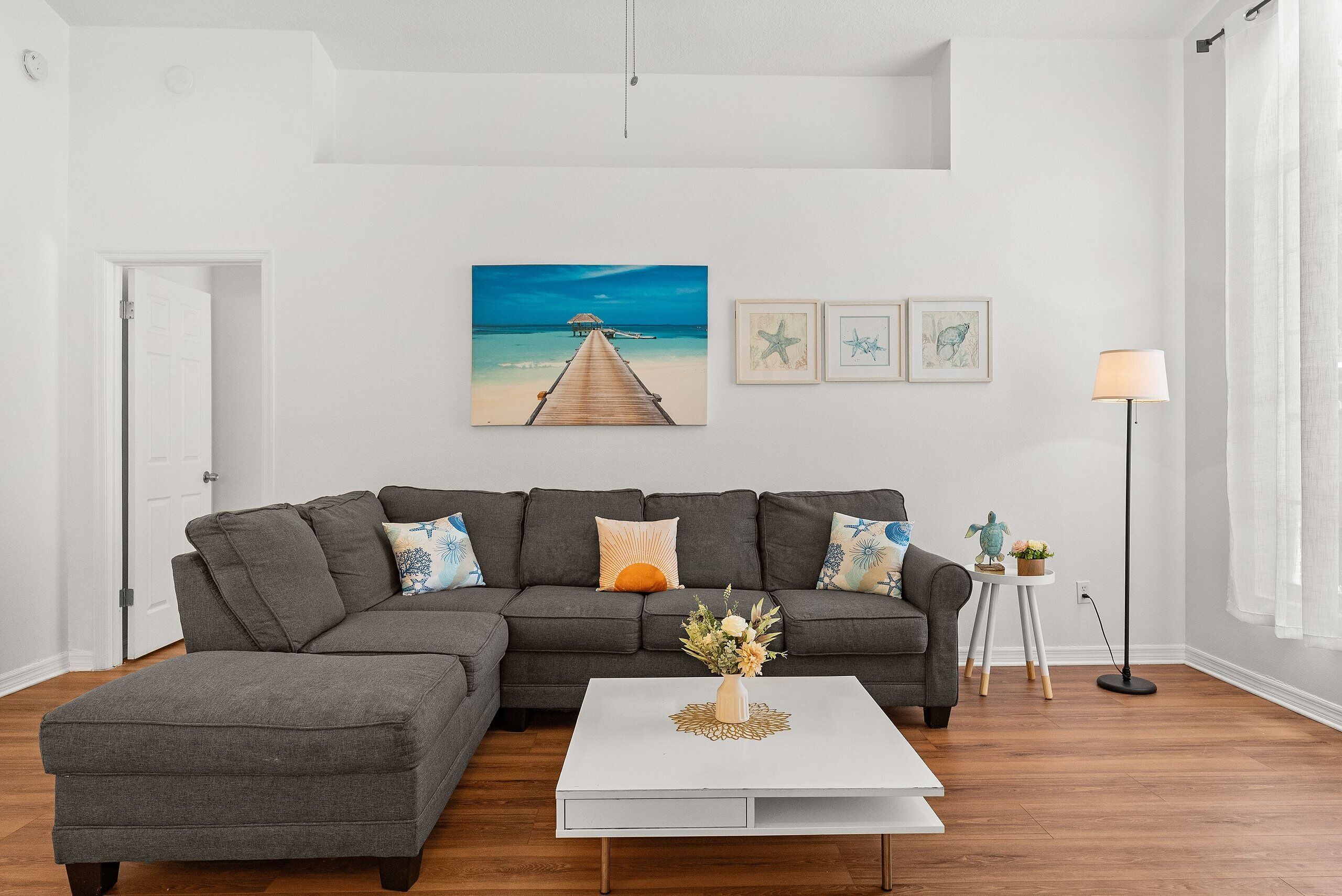 Un salon moderne avec un canapé d’angle gris, une table basse blanche et un tableau représentant une scène de plage.