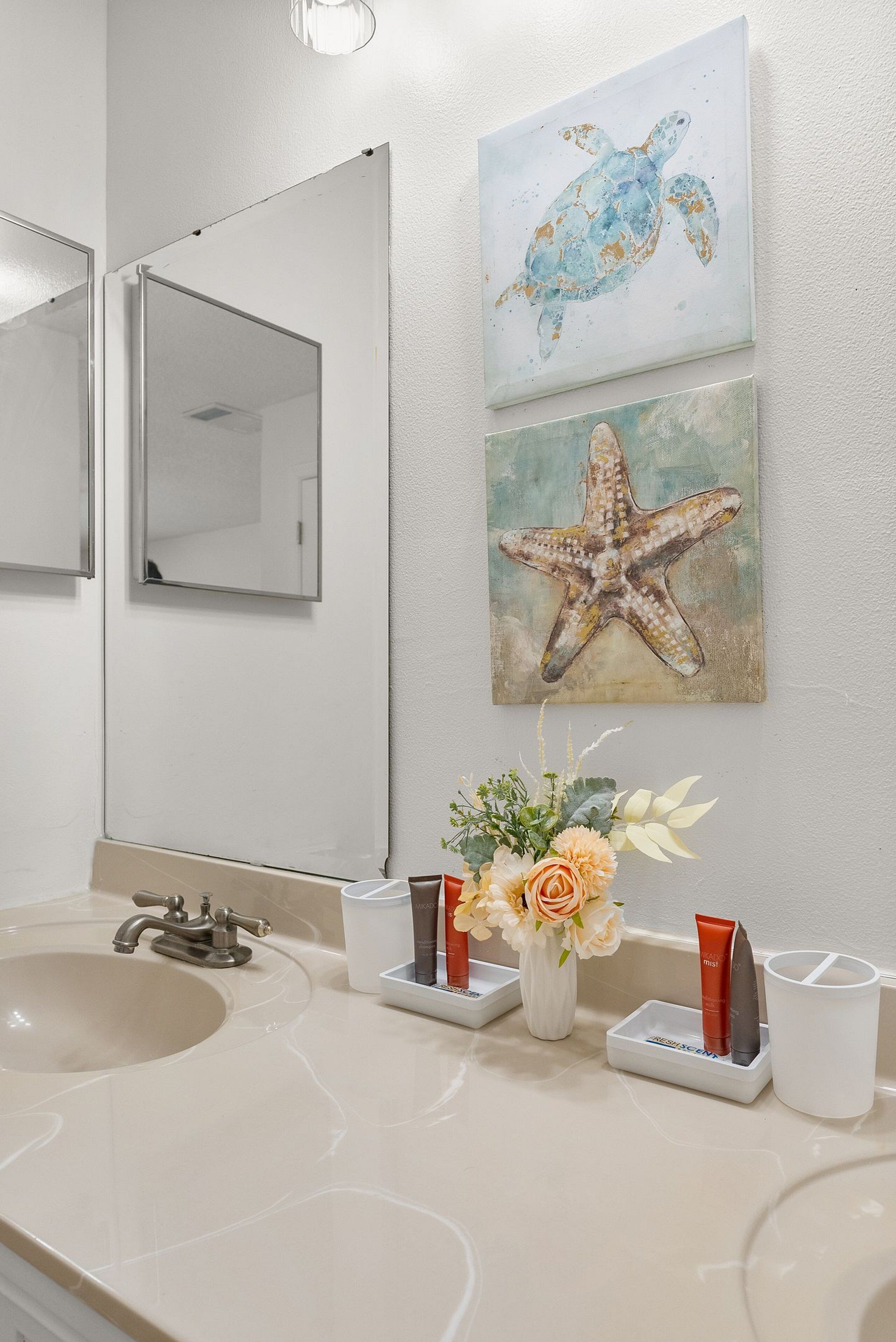 Une salle de bain avec un grand miroir, un lavabo et deux tableaux accrochés au mur représentant une tortue de mer et une étoile de mer.