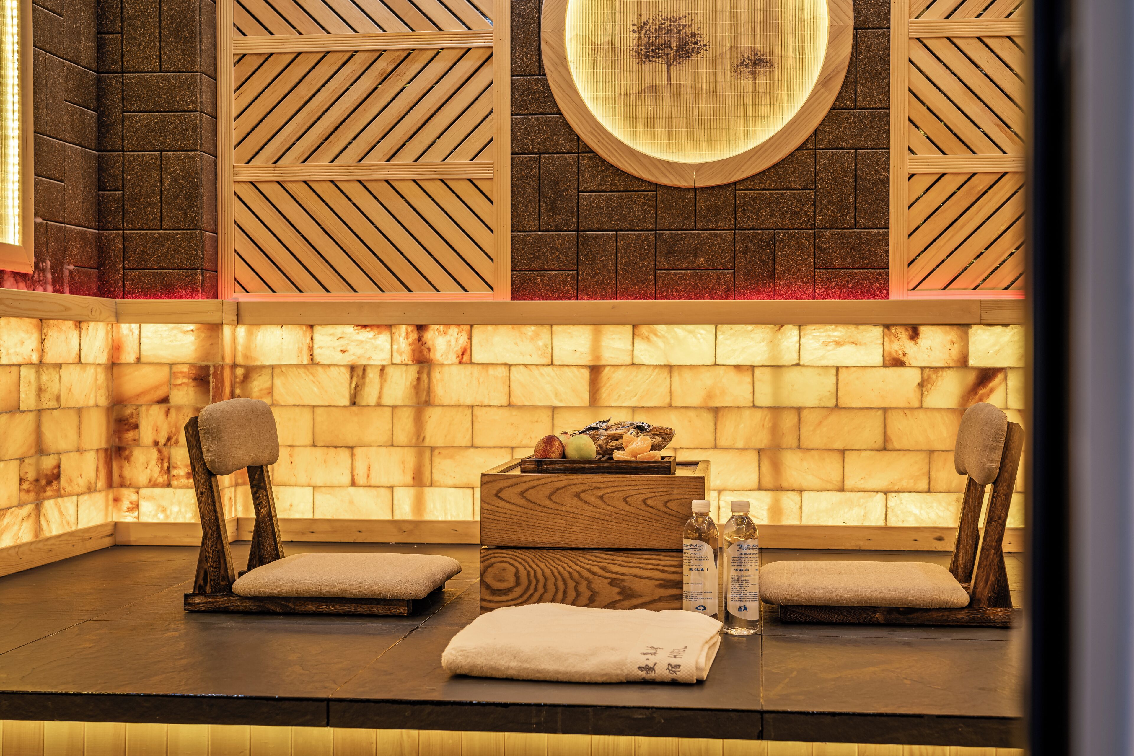 A spa room with two wooden chairs, a wooden bench, a small wooden table with fruits, bottles, and a towel.