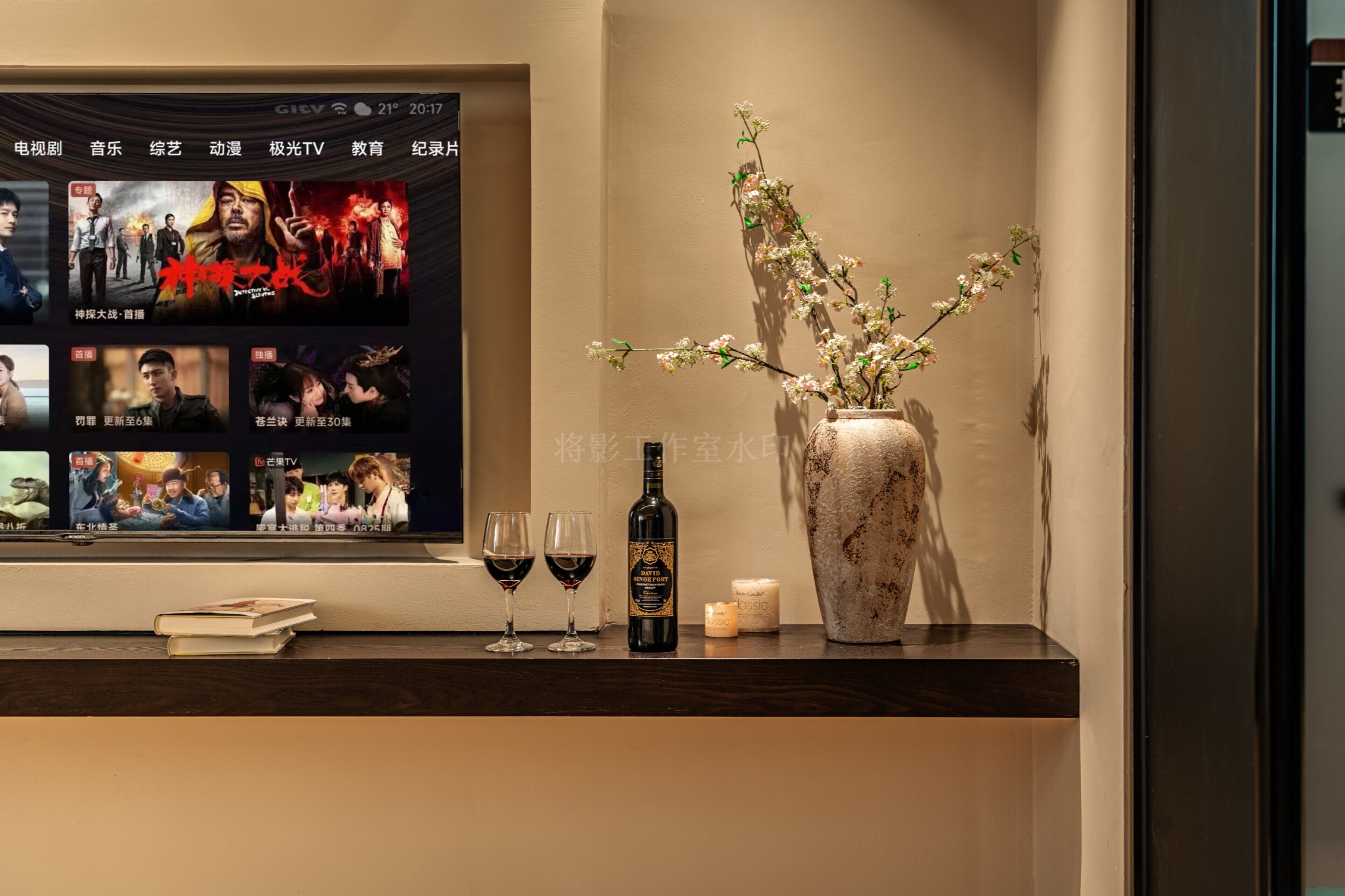 A hotel reception area with a TV displaying movie posters, a vase with flowers, wine glasses, a bottle of wine, and a candle on a counter.