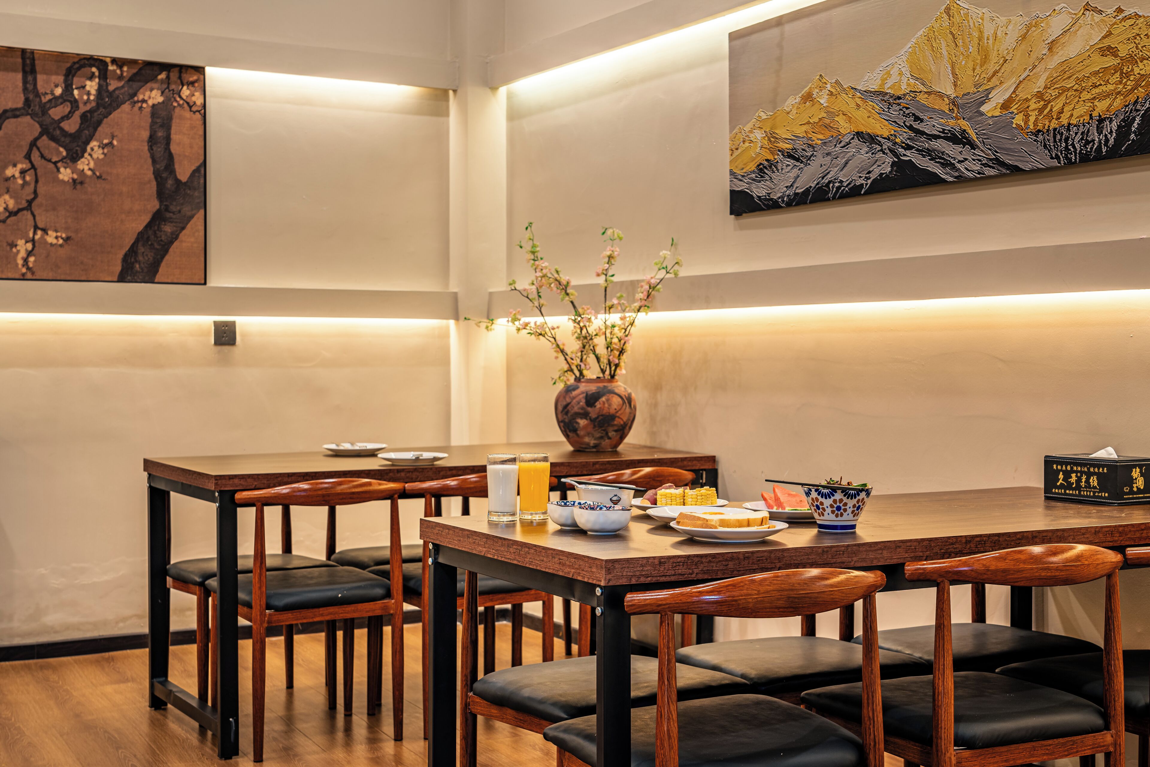 A dining area with wooden tables and chairs, a vase with flowers, and a painting on the wall.