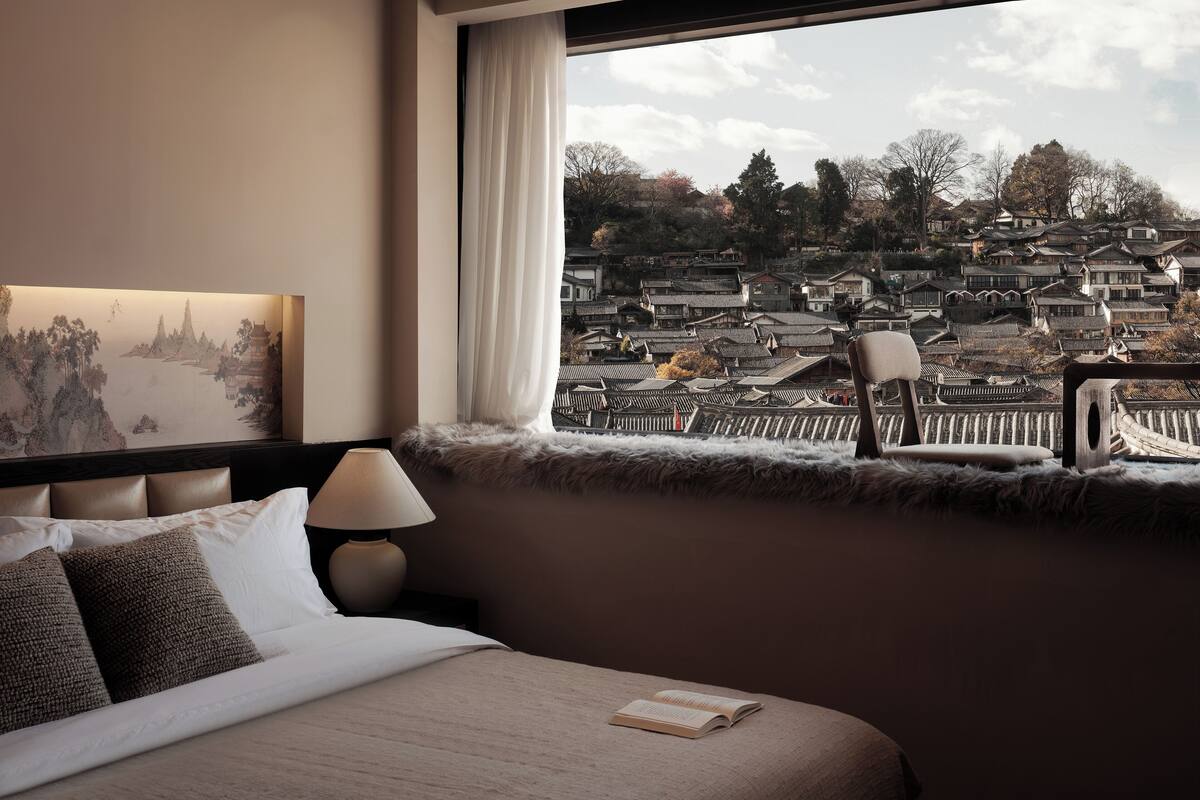 Ein Hotelzimmer mit einem Bett, einem Nachttisch mit Lampe, Blick auf traditionelle Gebäude durch ein Fenster und einem Sessel am Fenster.