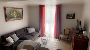 A living room with a sofa, a small round table, a chair, a TV on a wooden stand, and a painting on the wall.