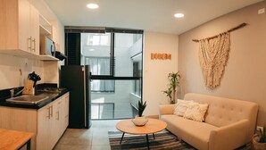 Eine moderne Küche mit einem schwarzen Kühlschrank, Holzschränken und einer Spüle. Ein runder Holztisch mit einer Pflanze in der Mitte. Ein beiges Sofa steht an der Wand, und ein Wandbehang aus Makramee ist sichtbar.