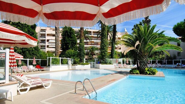 A swimming pool with lounge chairs and umbrellas, surrounded by palm trees and a building in the background.