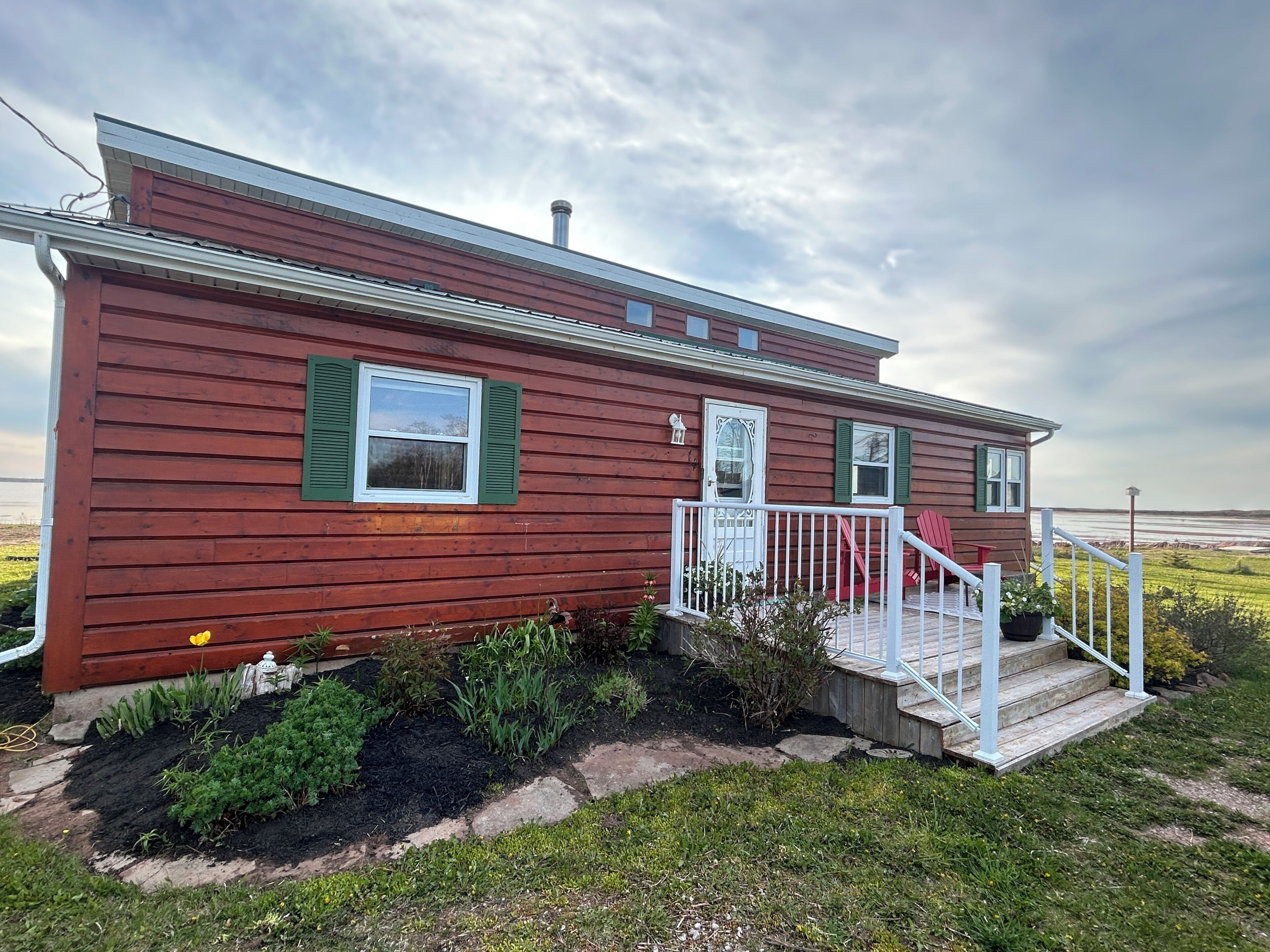 A single-story wooden house with a front porch, a small garden, and a view of the water.