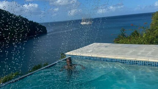 A clear blue pool with a person swimming, a boat in the distance, and a tropical landscape with a clear sky. - Poinsettia Villa (Marigot Bay)