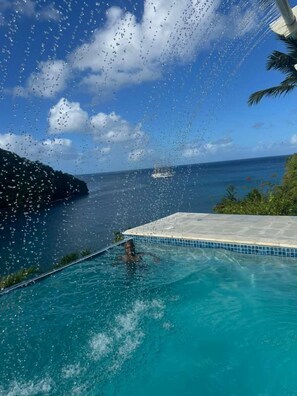 Uma piscina azul límpida com uma pessoa nadando, um barco ao longe e uma paisagem tropical com céu claro.