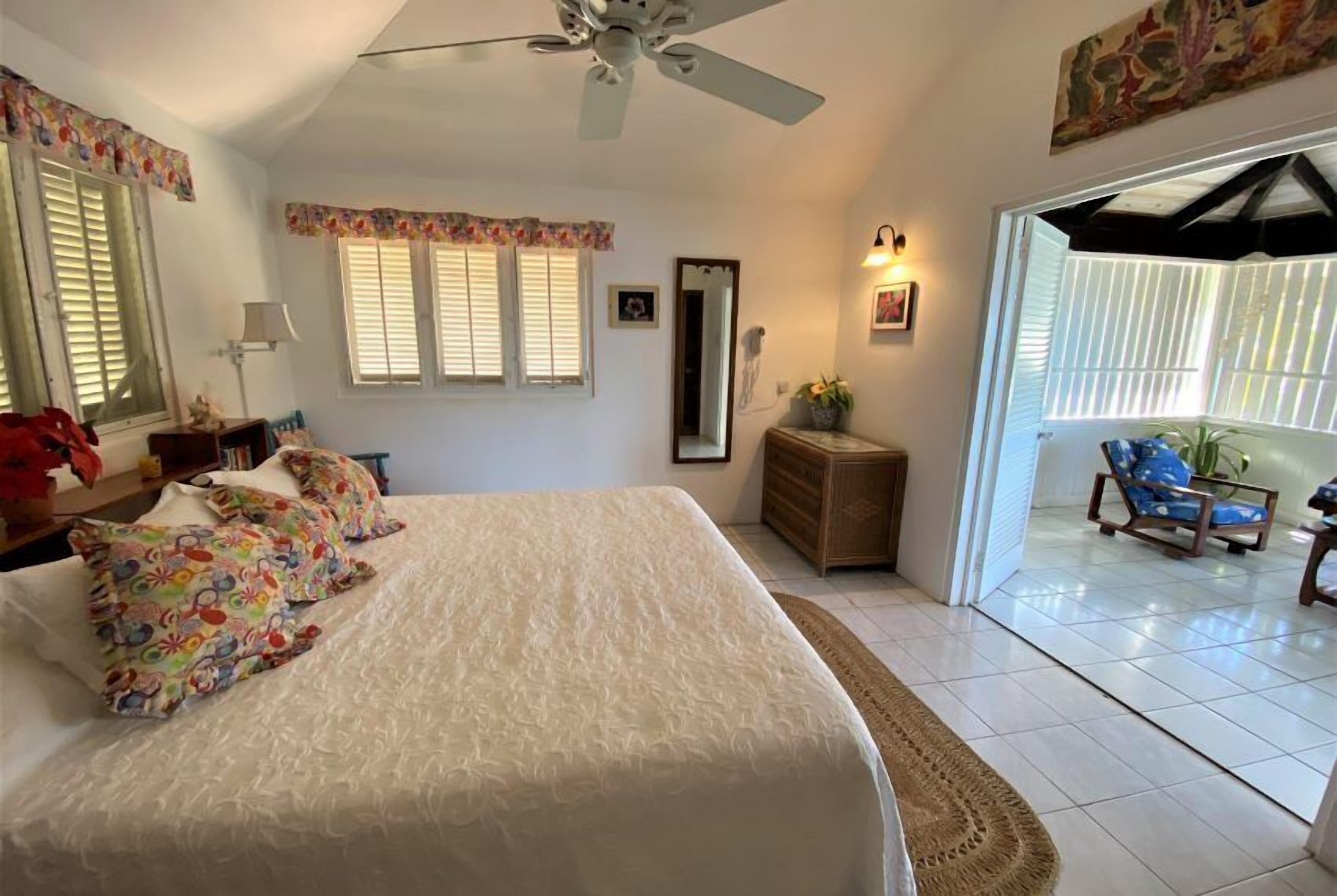 A bedroom with a bed, a ceiling fan, a dresser, a mirror, and a view of the outside.