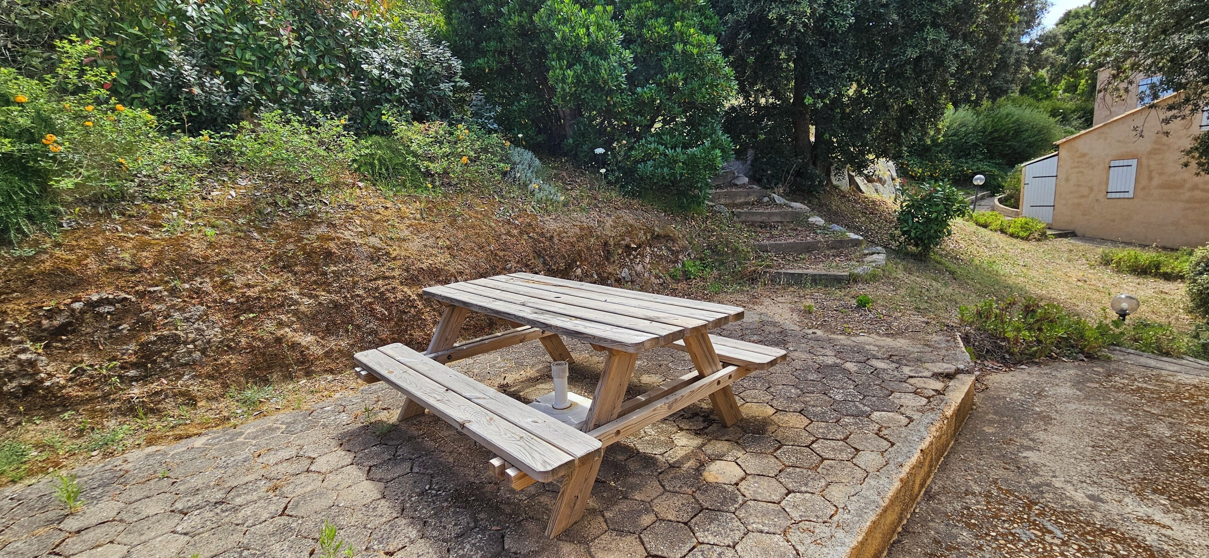 Ein hölzernes Picknicktisch auf einer sechseckigen Steinterrasse, umgeben von Grünpflanzen und einem Gebäude.