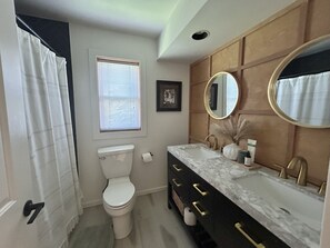 A bathroom with a toilet, a sink with a marble countertop, a vanity with gold handles, a mirror, and a window with blinds.