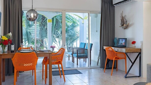 A modern living room with a dining area, featuring a glass door leading to a balcony with a view of palm trees.