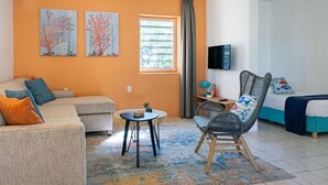 A modern living room with a sofa, a chair, a small table, and a bed. There is a window with curtains, a flat-screen TV mounted on the wall, and two paintings on the wall.