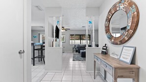 A modern interior with a white tiled floor, a wooden reception desk, a large decorative mirror, and a view into a living area with a sofa and a television. - Charming 3-bedroom house with WiFi, AC in Palm Harbor (Palm Harbor)