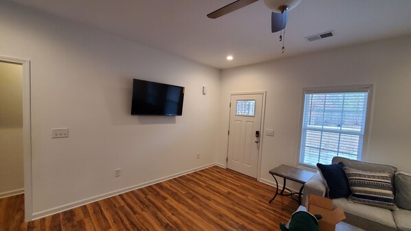 A living room with hardwood flooring, a flat-screen TV mounted on the wall, a door with a window, and a window with blinds.
