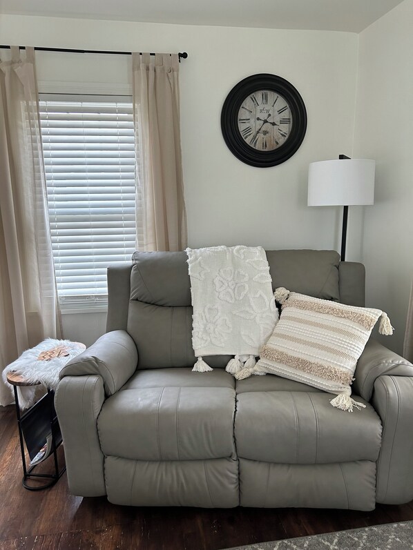 A living room with a grey sofa, a white throw blanket, a round wall clock, a floor lamp, and a small side table with a white furry throw. - Cottage in Sugarcreek with Hot Tub (Sugarcreek)