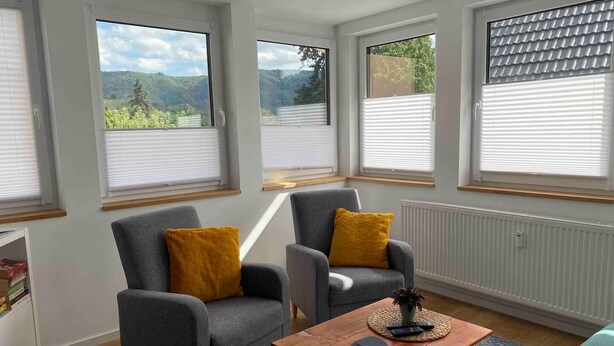 A modern living room with two grey armchairs, a wooden coffee table, and a view of the outdoors through large windows. - Sit back and relax with us between the Moselle and the vineyards. (Mehring)