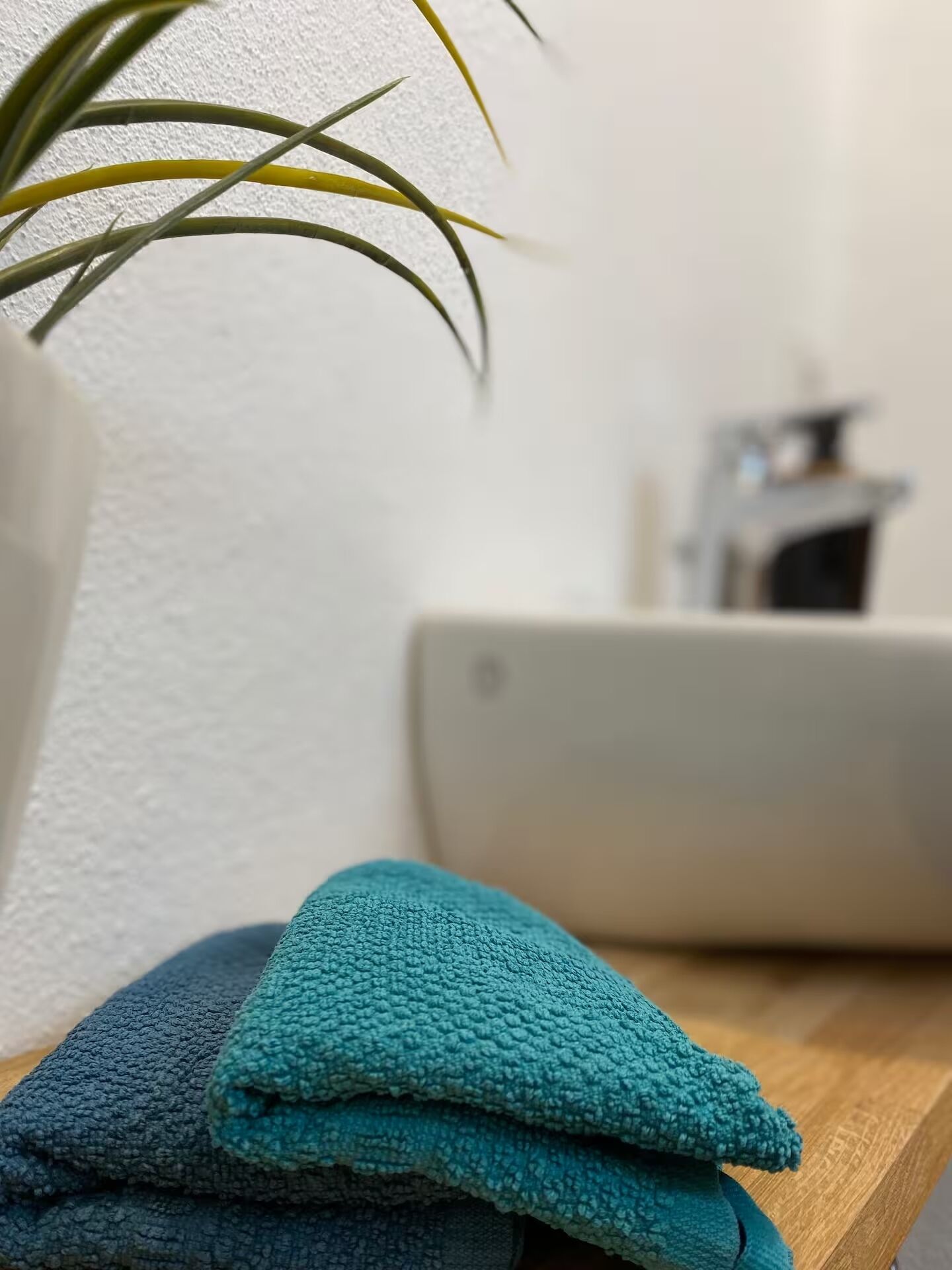 Ein Stapel gefalteter Handtücher auf einer Holztischplatte, eine Topfpflanze und ein unscharfer Hintergrund mit einer weißen Wand und einem Möbelstück.