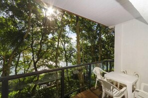 Un balcón con vistas a un bosque, una mesa blanca con sillas y una barandilla de cristal.