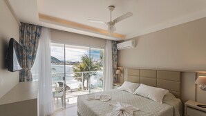 A hotel room with a bed, a ceiling fan, an air conditioning unit, a TV, and a balcony with a view of the sea and buildings. - Bombinhas Tourist (Bombinhas)