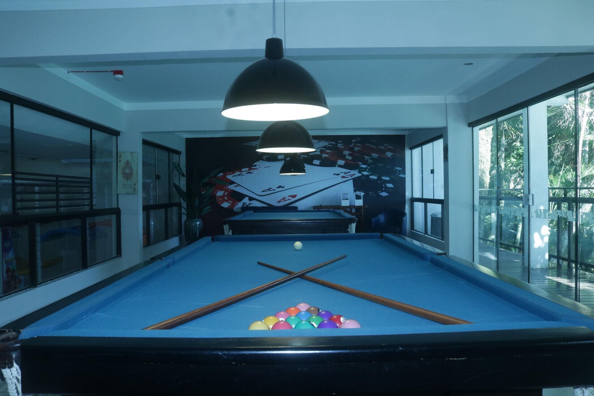 Una mesa de billar con taco y bolas, una lámpara colgante y una pared con un mural de cartas de juego.
