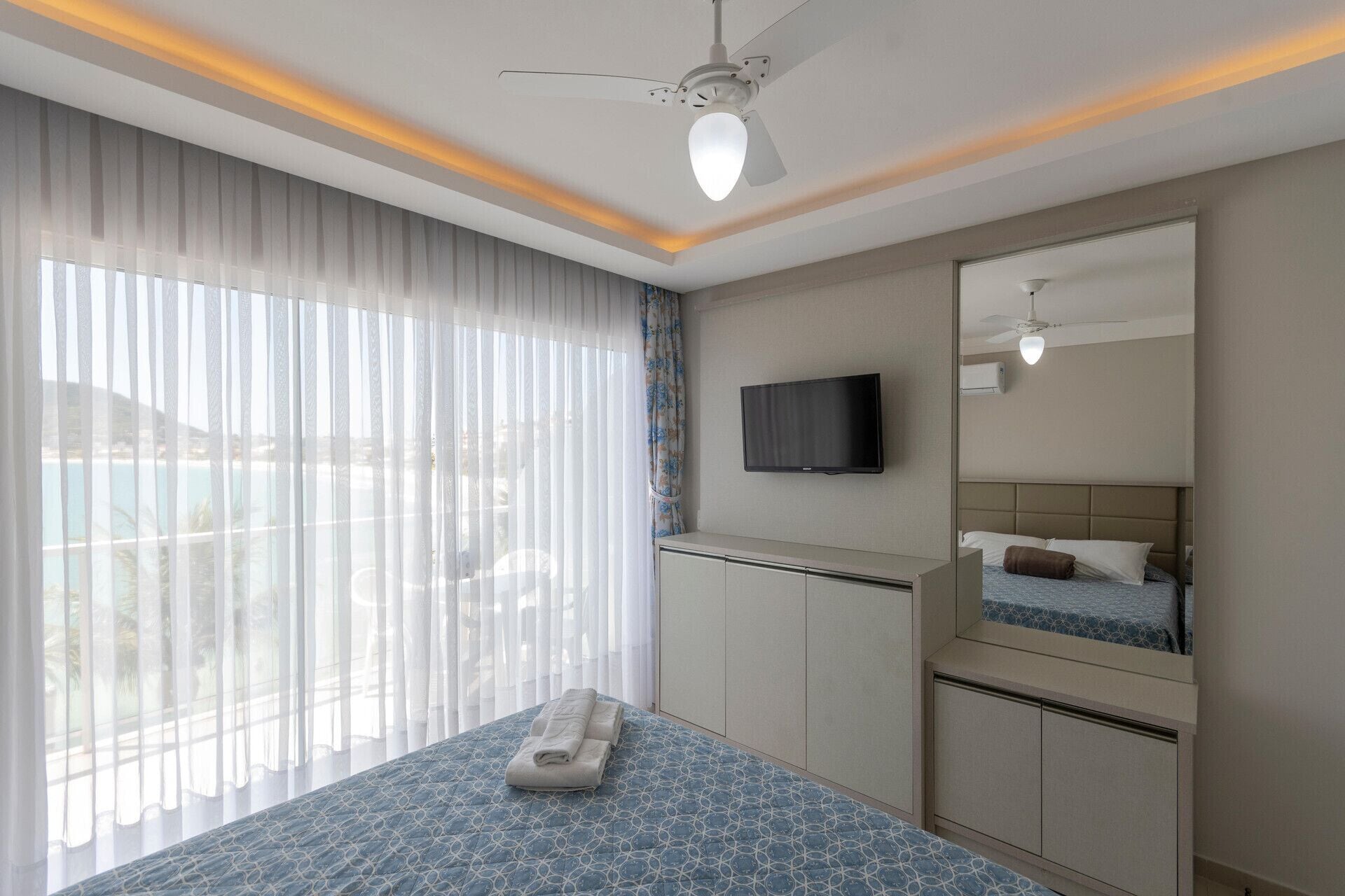 Habitación de hotel con cama, televisión, ventilador de techo y un gran ventanal con cortinas.