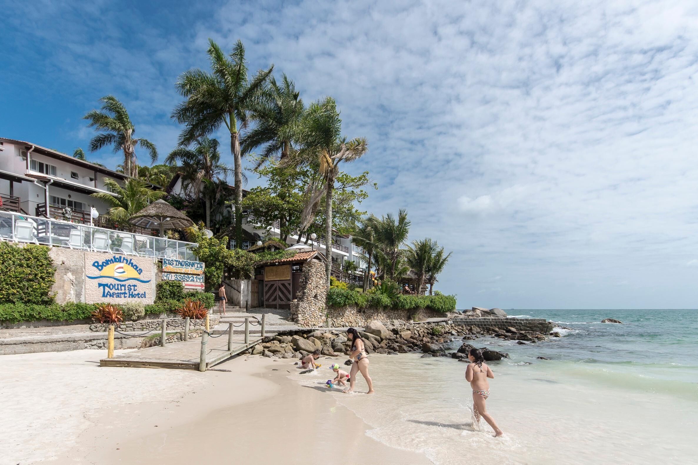 Uma praia com um letreiro do Bombiklas Tourist Art Hotel, palmeiras e uma costa rochosa.