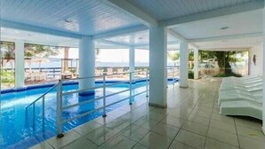 An indoor swimming pool with a view of the ocean, white poolside chairs, and a tiled floor. - Bombinhas Tourist (Bombinhas)