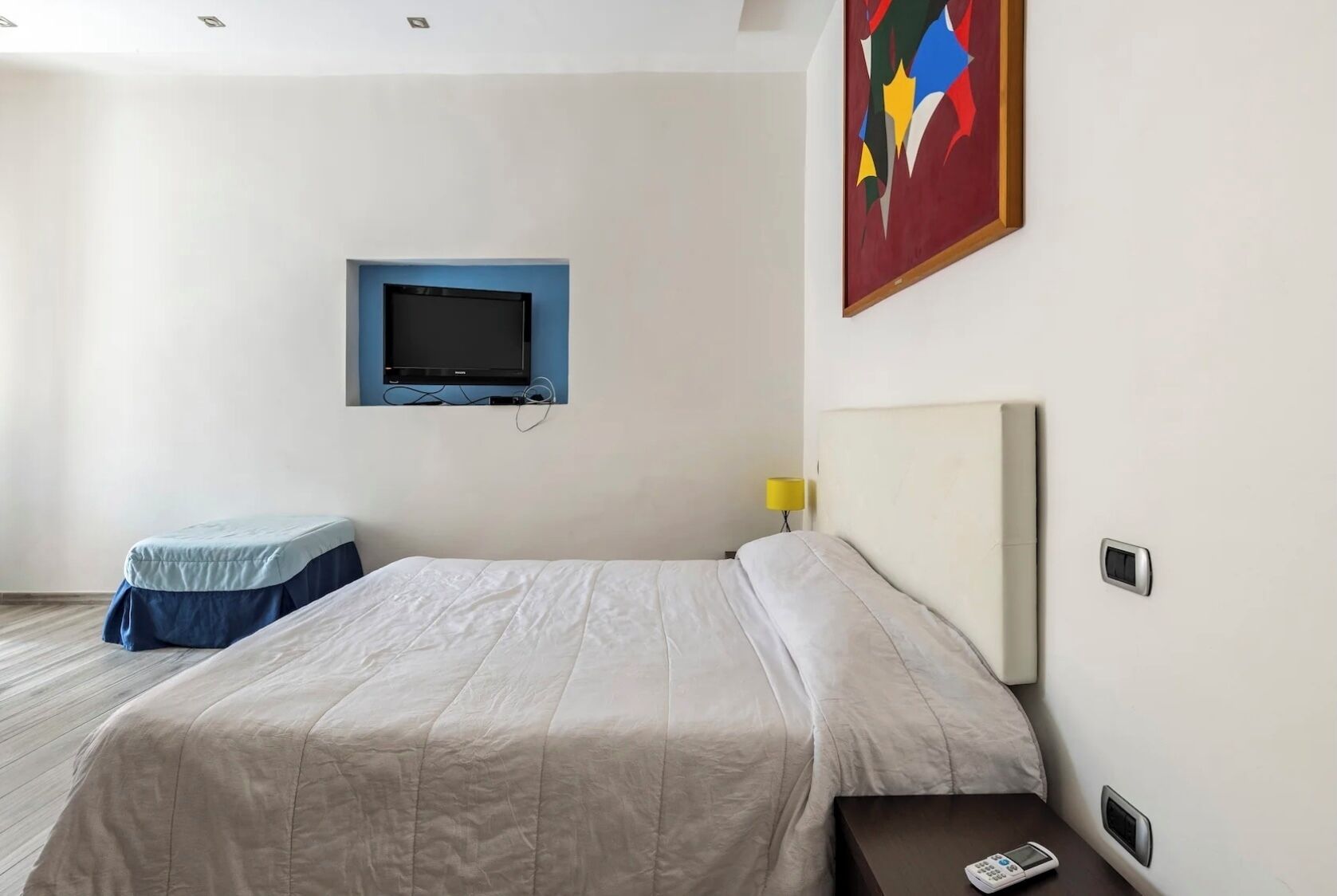 A hotel room with a bed, a television mounted on the wall, a bedside table with a lamp, and a painting on the wall.