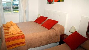 A hotel room with two beds, each with a beige bedspread and red pillows. There are towels on the beds and a window with curtains. - Hotel El Pescador (Mar de Ajo)