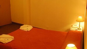 Ein kleines Zimmer mit einem Einzelbett, einer roten Bettdecke, einer Nachttischlampe und einem auf dem Bett liegenden Handtuch.
