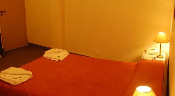 Une petite chambre avec un lit simple, une couverture rouge, une lampe de chevet et une serviette pliée posée sur le lit.