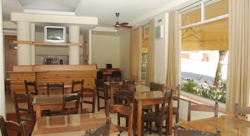 Un comedor con mesas y sillas de madera, una zona de bar con televisor, y grandes ventanales con vistas al exterior.