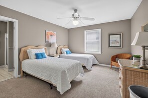 Quarto com duas camas, um ventilador de teto, uma janela com persianas, uma cadeira de vime e uma cômoda.