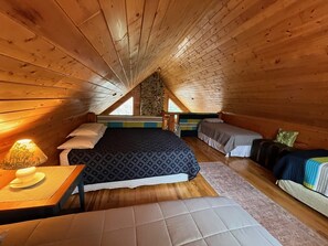 Un dormitorio acogedor en el ático, con techo inclinado, una cama grande, una cama más pequeña, una mesita de noche con lámpara y un banco.