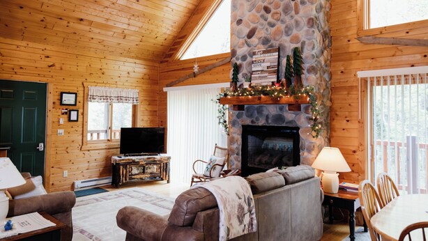 A cozy cabin interior with a stone fireplace, wooden walls, a television, and a dining area.