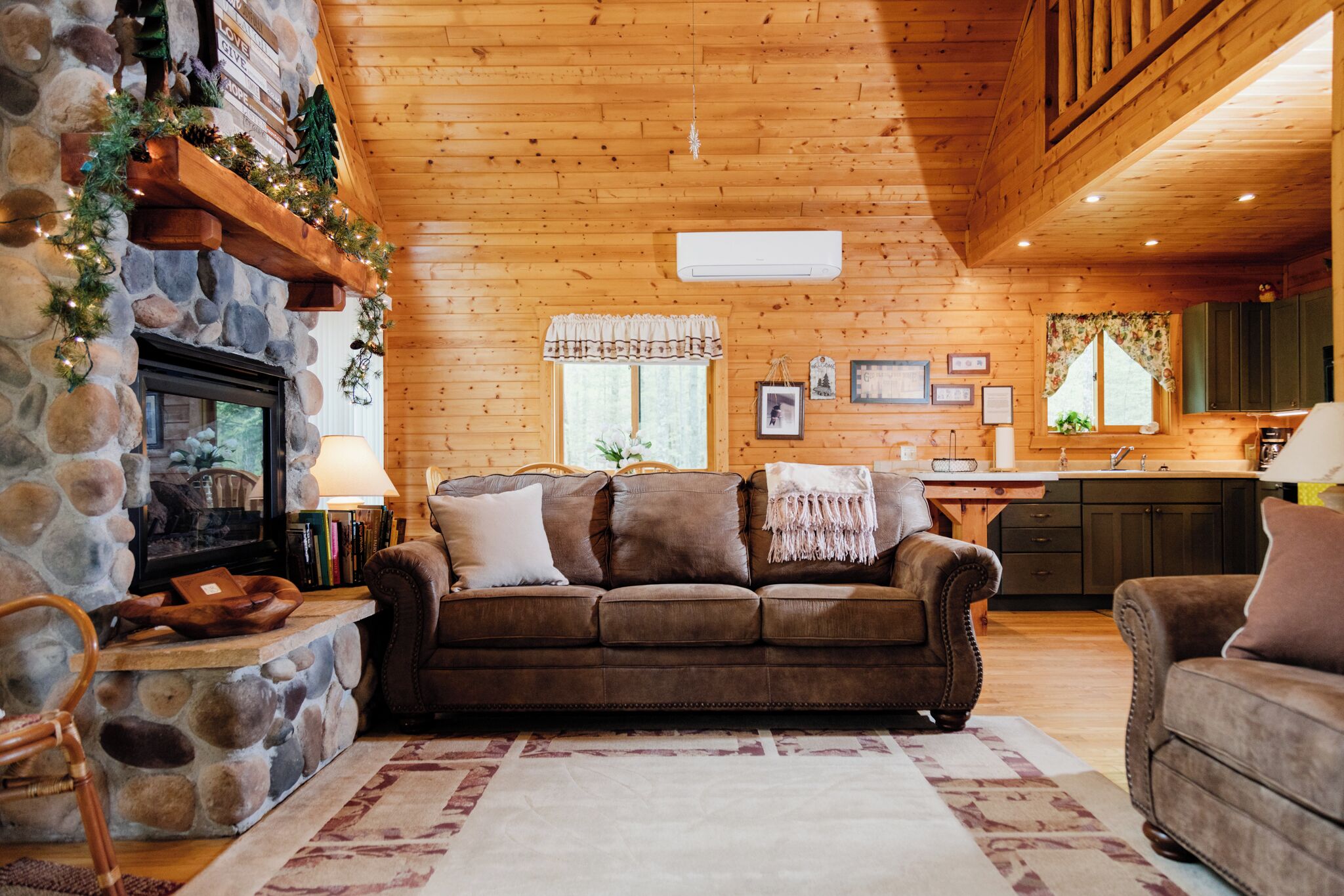 A cozy log cabin interior with a stone fireplace, wooden walls, a brown leather sofa, and a kitchen area.
